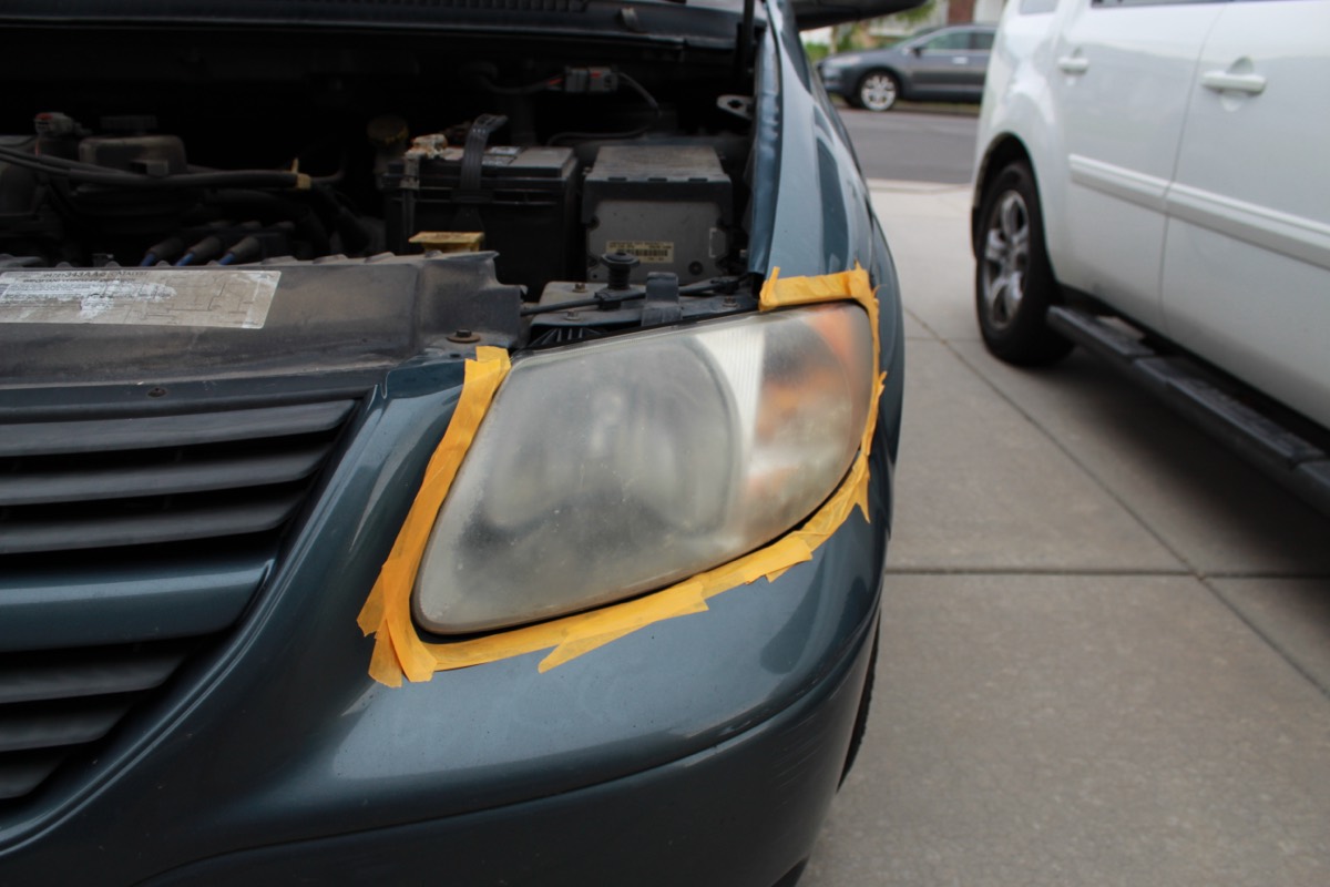 Heavily oxidized, foggy headlight on a green Dodge Grand Caravan, masked off with tape before restoration