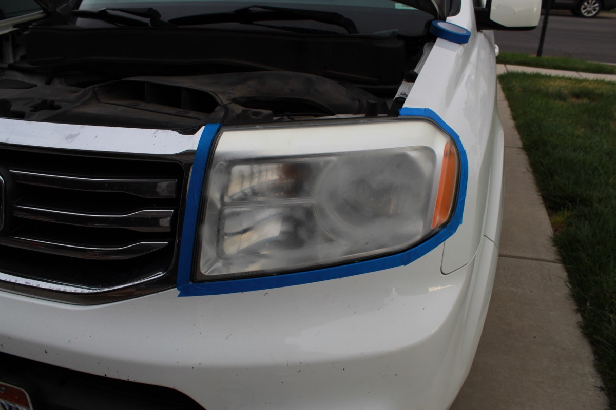 Driver side headlight of a white Honda Pilot, foggy and masked with blue tape before work begins